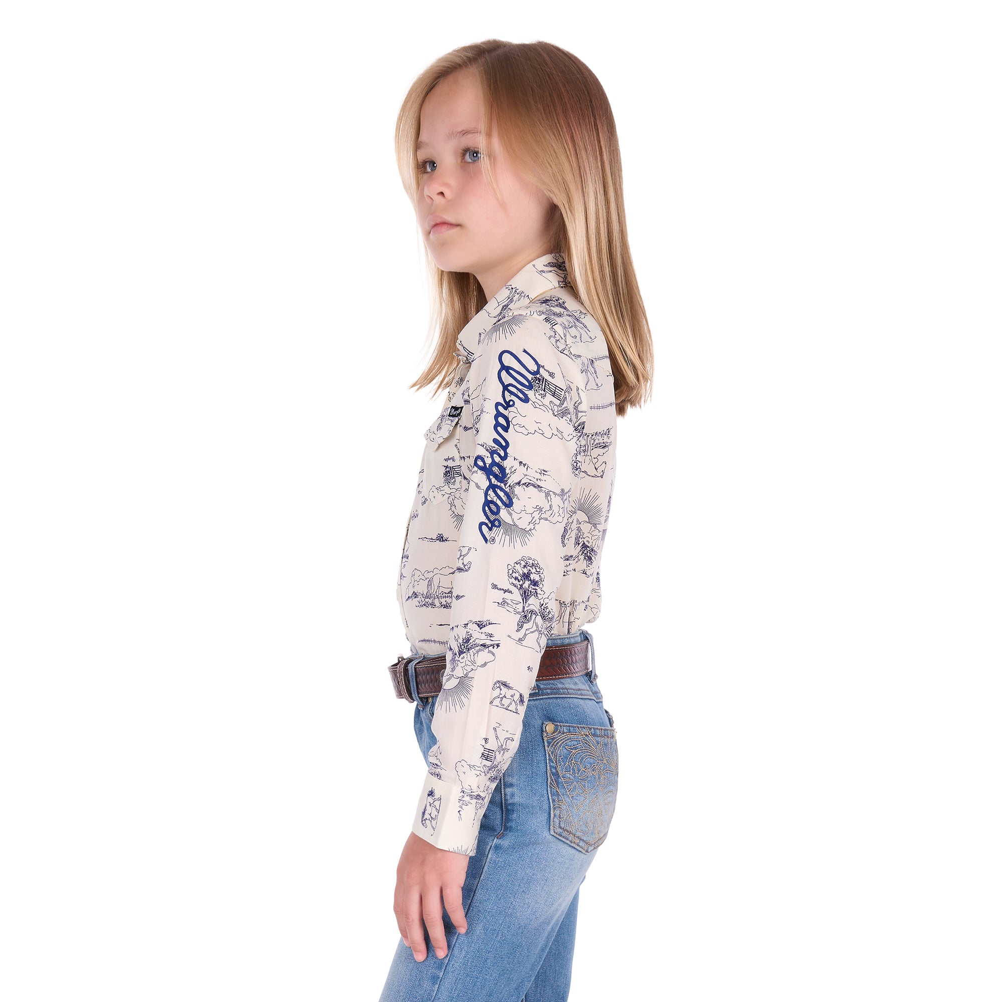A young girl with blonde hair stands in profile against a white background wearing a Wrangler Girls Ivory Logo Long Sleeve Shirt (X6S5127602) with a horse print on a cream base, paired with blue jeans and a dark tan Wrangler Children's Denny Belt (XCP7951BLT).