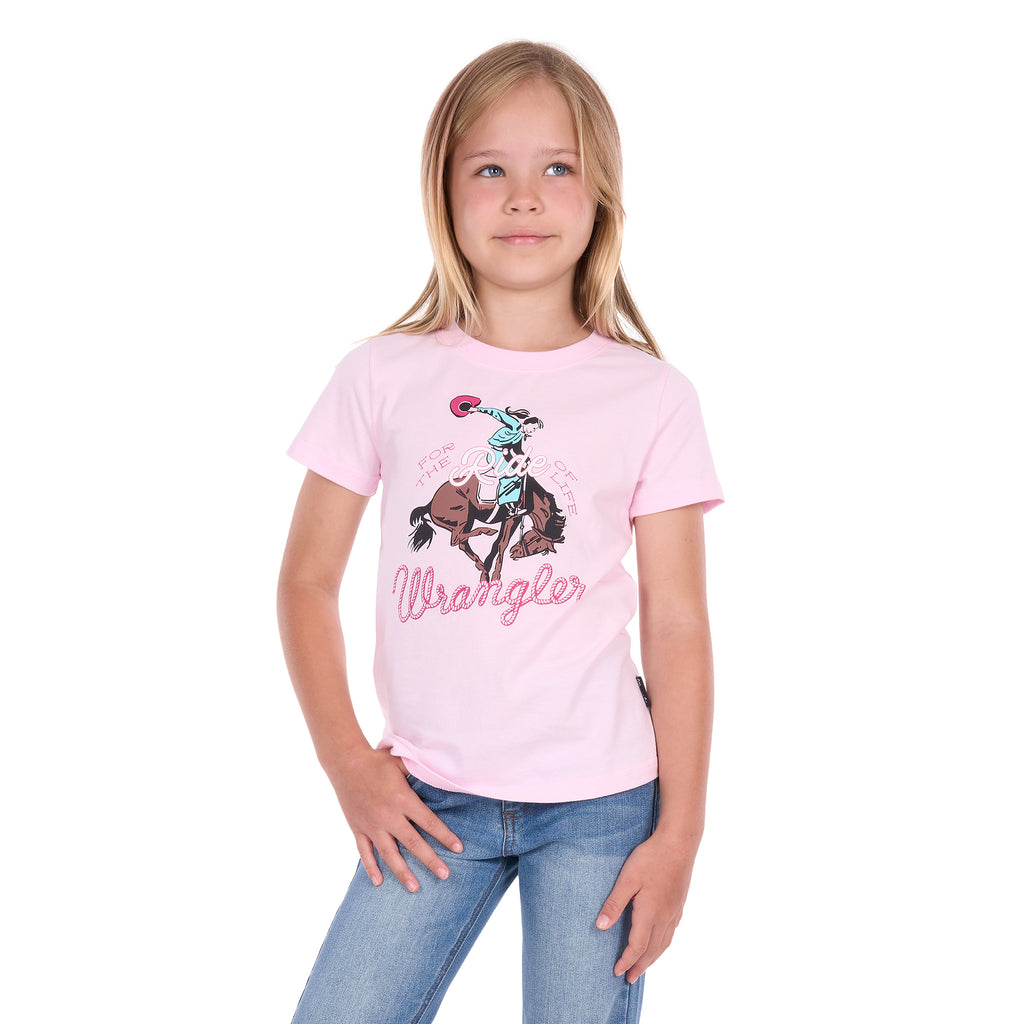 A young girl with blonde hair wearing a pastel pink Wrangler Girls Jenna T-Shirt (X6S5598639) with a cowboy graphic and blue jeans, standing against a plain white background.