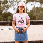 A young girl wearing a pastel pink  Wrangler Girls Jenna T-Shirt (X6S5598639) with a cowboy graphic and a Wrangler rope logo, paired with a denim mini skirt and a pink Wrangler Children's Imogen Full Back Cap (X6S5947CAP), standing outdoors in front of a wooden fence.