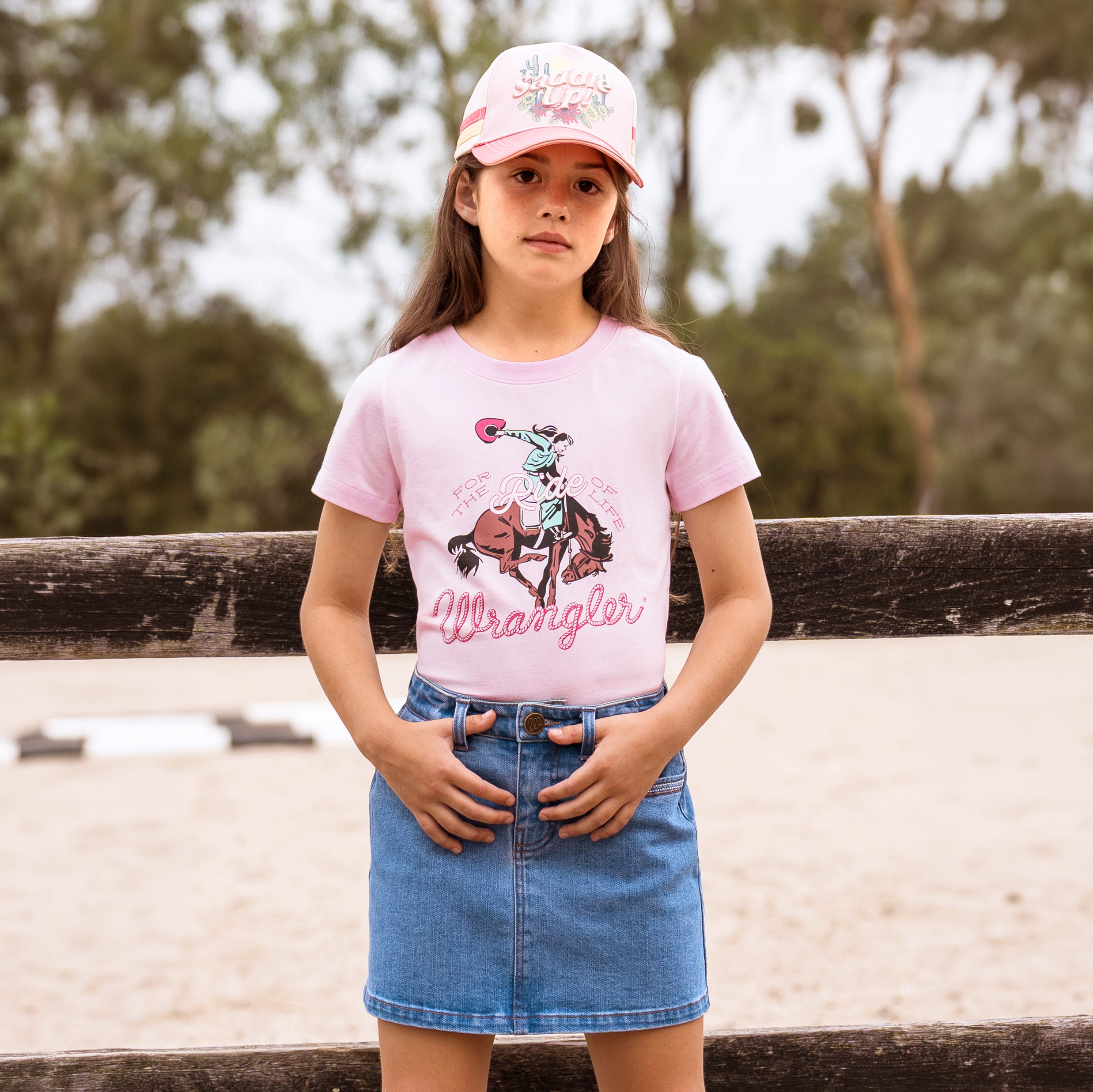 A young girl wearing a pastel pink  Wrangler Girls Jenna T-Shirt (X6S5598639) with a cowboy graphic and a Wrangler rope logo, paired with a denim mini skirt and a pink Wrangler Children's Imogen Full Back Cap (X6S5947CAP), standing outdoors in front of a wooden fence.