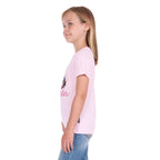 A young girl in profile with blonde hair wearing a pastel pink Wrangler Girls Jenna T-Shirt (X6S5598639) with a cowboy graphic and blue jeans, standing against a plain white background.