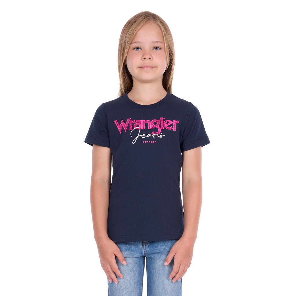 A young girl with blonde hair wearing a navy blue Wrangler Girls Maya T-Shirt (X6S5598649) and blue jeans, standing against a plain white background.