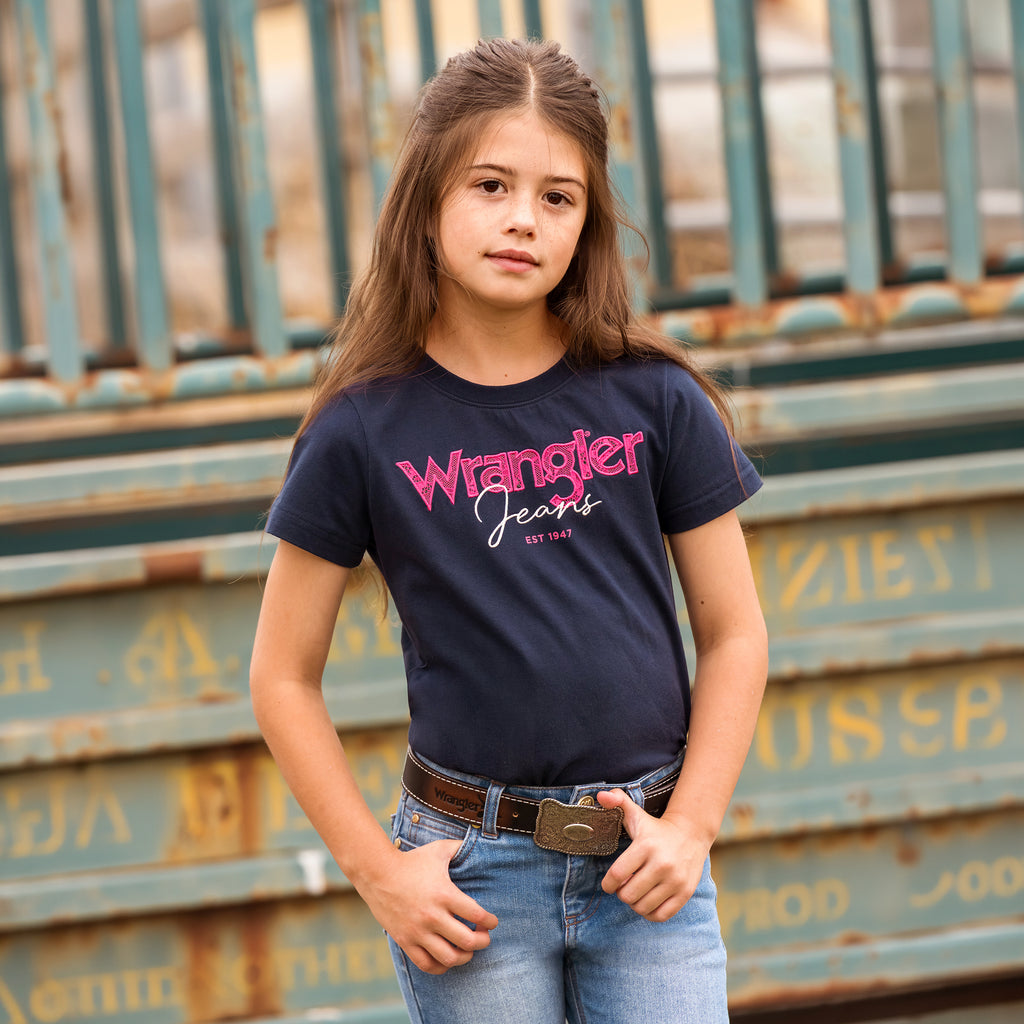 A young girl with long brown hair stands in front of a weathered teal metal structure, wearing a navy blue Wrangler Girls Maya Tee (X6S5598649) Childrens, an Abrasion Logo Belt (XCP3902BEL) and Wrangler Girls Lucy Bootcut Jeans (XCP5201719) in faded blue.