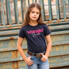 A young girl with long brown hair stands in front of a weathered teal metal structure, wearing a navy blue Wrangler Girls Maya Tee (X6S5598649) Childrens, an Abrasion Logo Belt (XCP3902BEL) and Wrangler Girls Lucy Bootcut Jeans (XCP5201719) in faded blue.