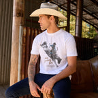 A man wearing a white Wrangler Men's Michael Short Sleeve graphic T-Shirt (X6S1557684) t-shirt, dark blue jeans, and a Wrangler Hudson Bangora Hat (XCP1928HAT) cowboy hat, sitting on a brown leather couch in a rustic barn setting.