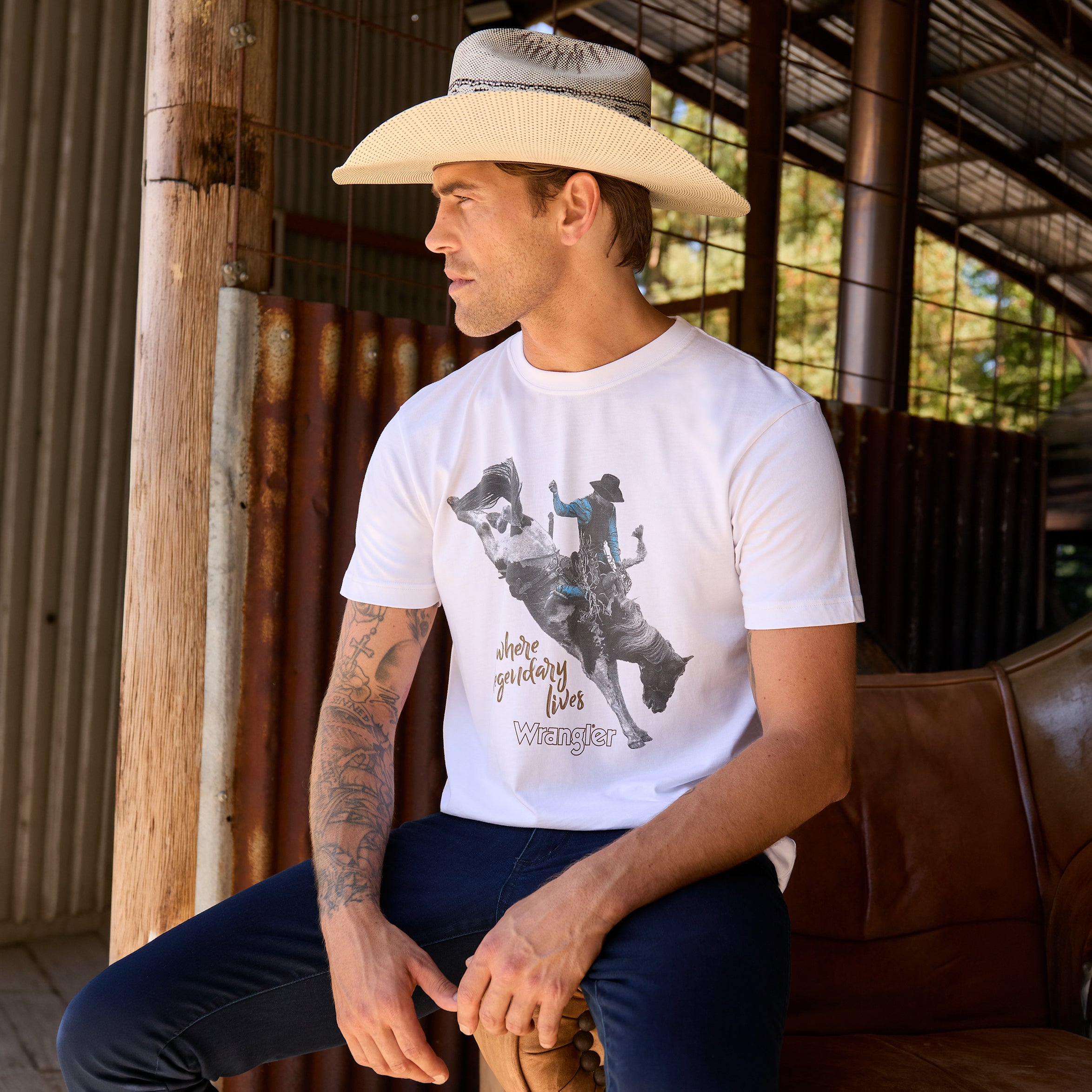A man wearing a white Wrangler Men's Michael Short Sleeve graphic T-Shirt (X6S1557684) t-shirt, dark blue jeans, and a Wrangler Hudson Bangora Hat (XCP1928HAT) cowboy hat, sitting on a brown leather couch in a rustic barn setting.