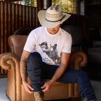 A man wearing a white Wrangler Men's Michael Short Sleeve graphic T-Shirt (X6S1557684) t-shirt, dark blue jeans, and a Wrangler Hudson Bangora Hat (XCP1928HAT) cowboy hat, sitting on a brown leather couch in a rustic barn setting.