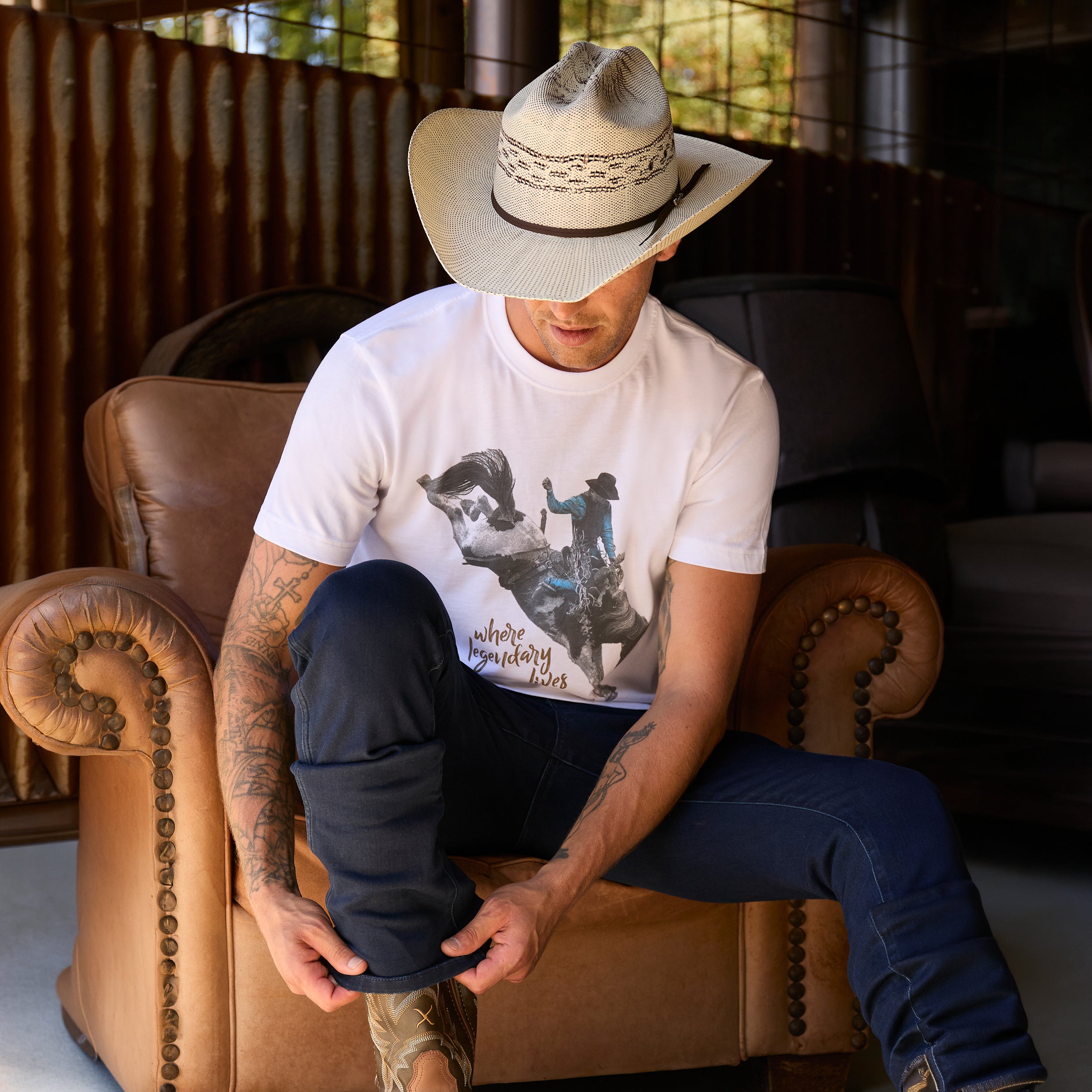 A man wearing a white Wrangler Men's Michael Short Sleeve graphic T-Shirt (X6S1557684) t-shirt, dark blue jeans, and a Wrangler Hudson Bangora Hat (XCP1928HAT) cowboy hat, sitting on a brown leather couch in a rustic barn setting.