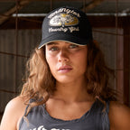 A woman wearing a Wrangler Jasmine Trucker Cap (X6S2938CAP) in Charcoal with a metallic gold and white embroidered steer skull graphic on the front panel, Wrangler logo embroidery and white embroidered script text reading 'Wrangler Country Girl'. Also wearing a Wrangler Womens Jasmine Tank (X6S2508648) in Charcoal, photographed in a rustic shed.