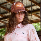A woman wearing a Wrangler Jenna High Profile Ponytail Trucker Cap (X6S2939CAP) in Copper and a Wrangler Women's Hazel Logo Long Sleeved Shirt (XCP2111608) in Blush, posed in a rustic structure.