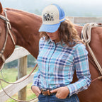 A light blue and cream Wrangler Josie High Profile Trucker Cap (X6S2943CAP) featuring a western-themed graphic print on the front panel and cream embroidered text on the brim, photographed on a model wearing a teal and pink Wrangler Women's Luisa Shirt (X6S2100607) standing next to a horse with western tack.