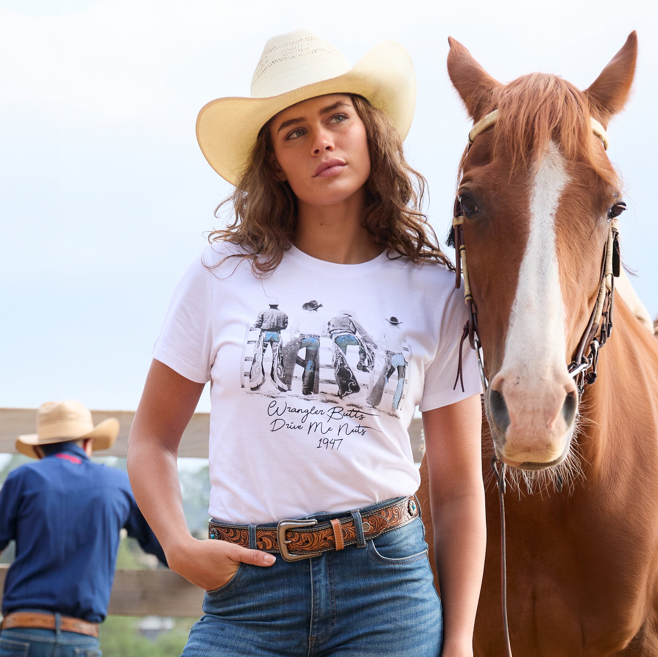 A woman modelling a white graphic Wrangler Women's Josie Short Sleeve T-Shirt (XCP2598641) with a black and white print of riders on a fence and the text 'Wrangler Butts Drive Me Nuts' on the front, paired with mid indigo Wrangler Etta Bootcut Jeans Bailey (XCP2255711), a tan Wrangler Siara Belt (X6S2916BLT), and a natural Wrangler Jhonson Bangora Cowboy Hat (X4Y2025JHO) standing next to a brown horse outdoors.