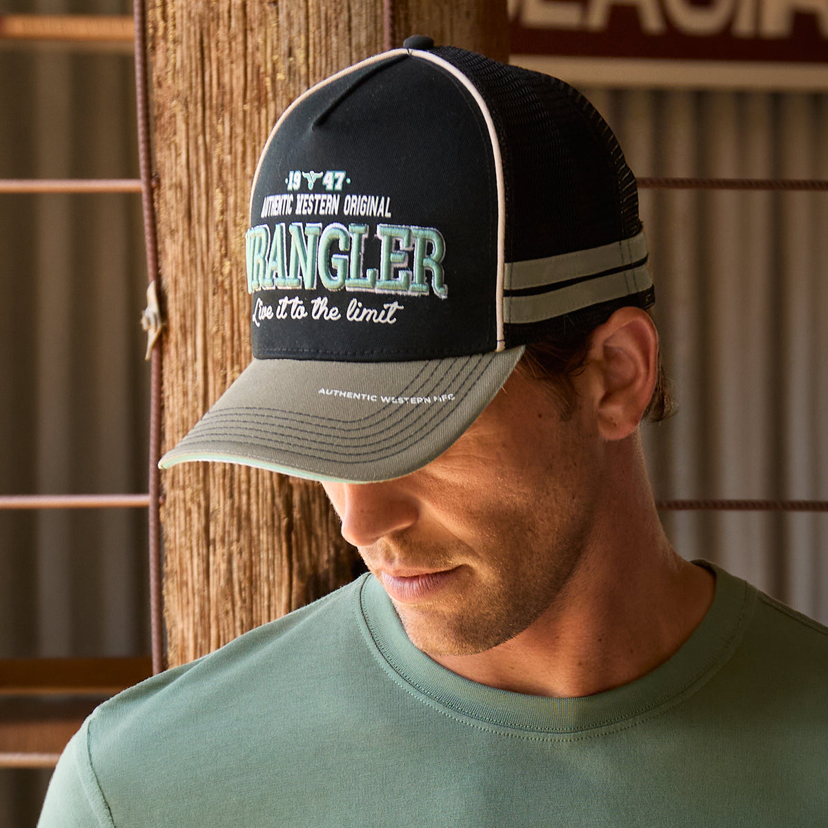 A man wearing a black and sage green Wrangler Layton High Profile Trucker Cap (X6S1945CAP) featuring a black front panel with white and teal Wrangler print and embroidery on the front, black mesh back panels, with a curved brim and side panel taping in sage green paired with a sage green t-shirt in a rustic shed setting.
