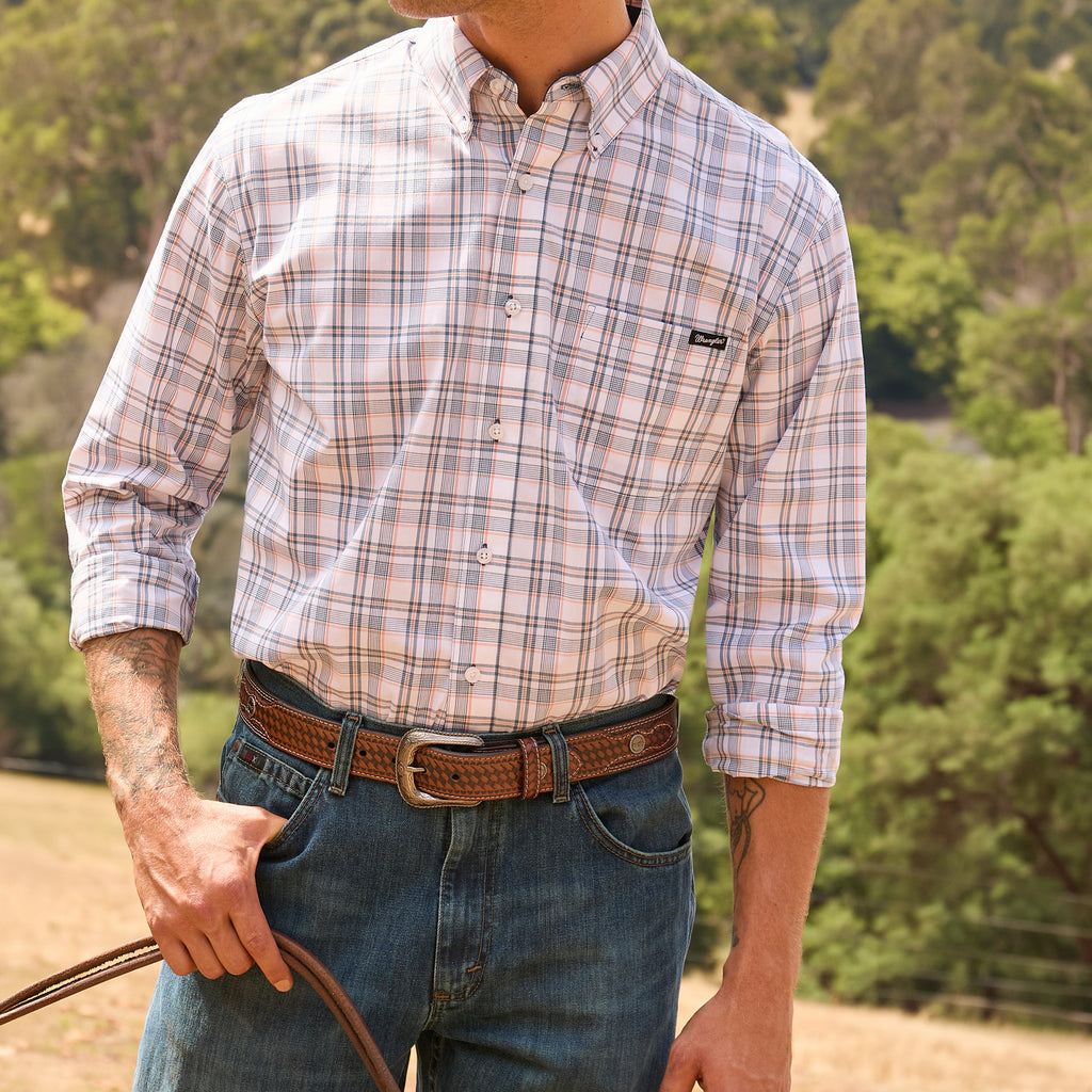 A man wearing a long-sleeve button-down plaid shirt in pink, beige, and white tones, tucked into blue jeans with a tan Wrangler Leonard Belt (X6S1909BLT), standing outdoors against a blurred green forest background.