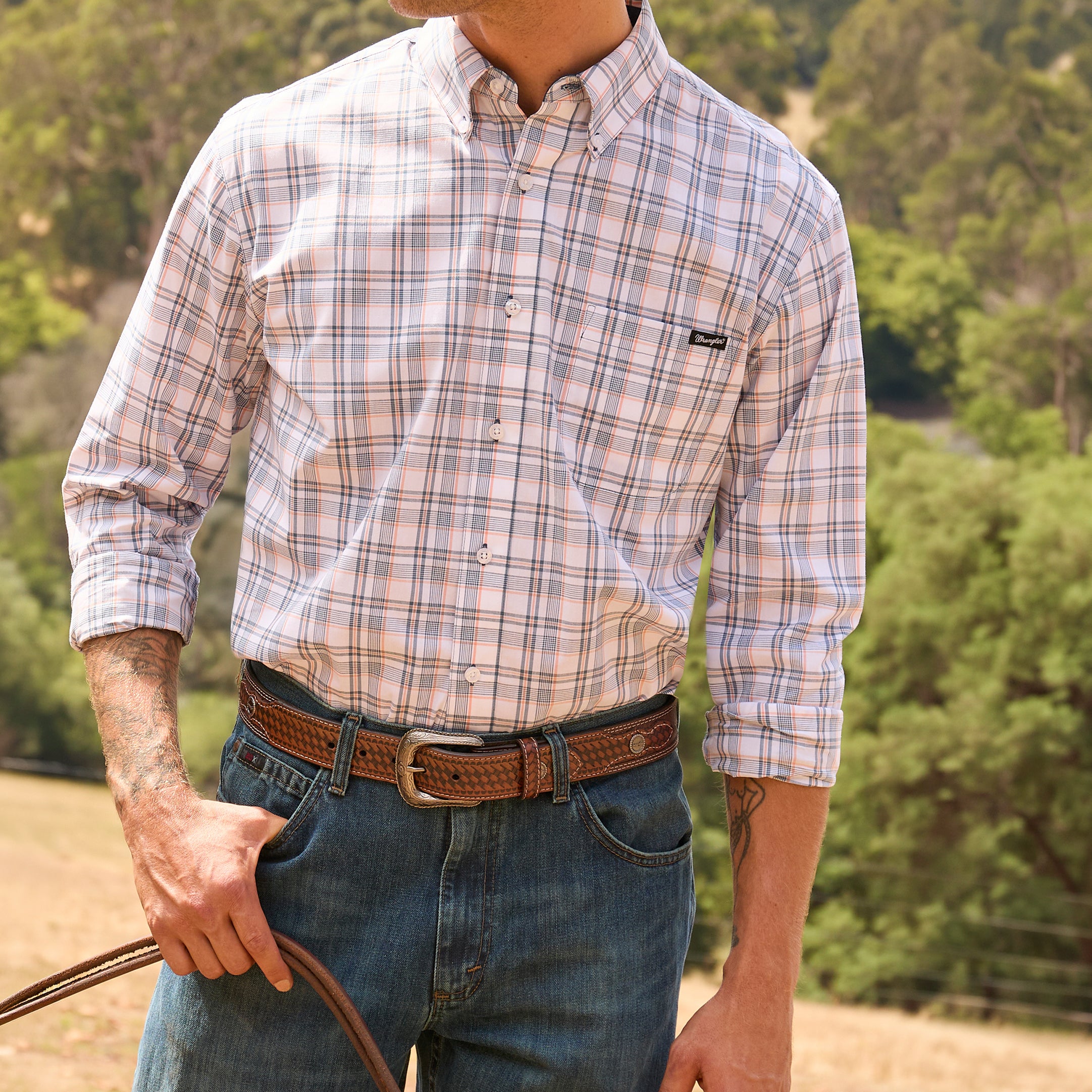A man wearing a long-sleeve button-down plaid shirt in pink, beige, and white tones, tucked into blue jeans with a tan Wrangler Leonard Belt (X6S1909BLT), standing outdoors against a blurred green forest background.