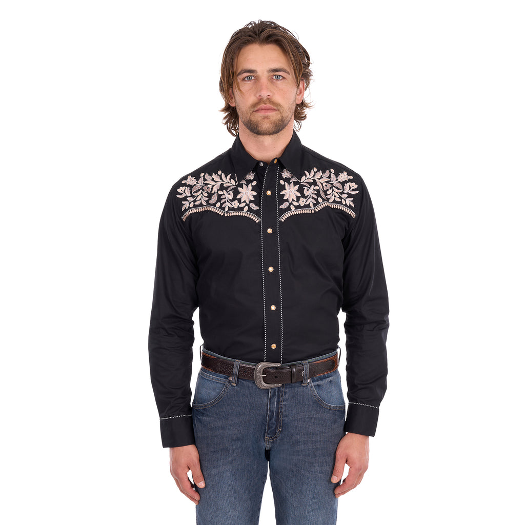 A man wearing a black Wrangler Men's Anthony Embroidered L/S Shirt (X6S1113674) Black with intricate white floral embroidery across the chest, paired with blue jeans and a brown belt, against a plain white background.
