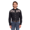 A man wearing a black Wrangler Men's Anthony Embroidered L/S Shirt (X6S1113674) Black with intricate white floral embroidery across the chest, paired with blue jeans and a brown belt, against a plain white background.