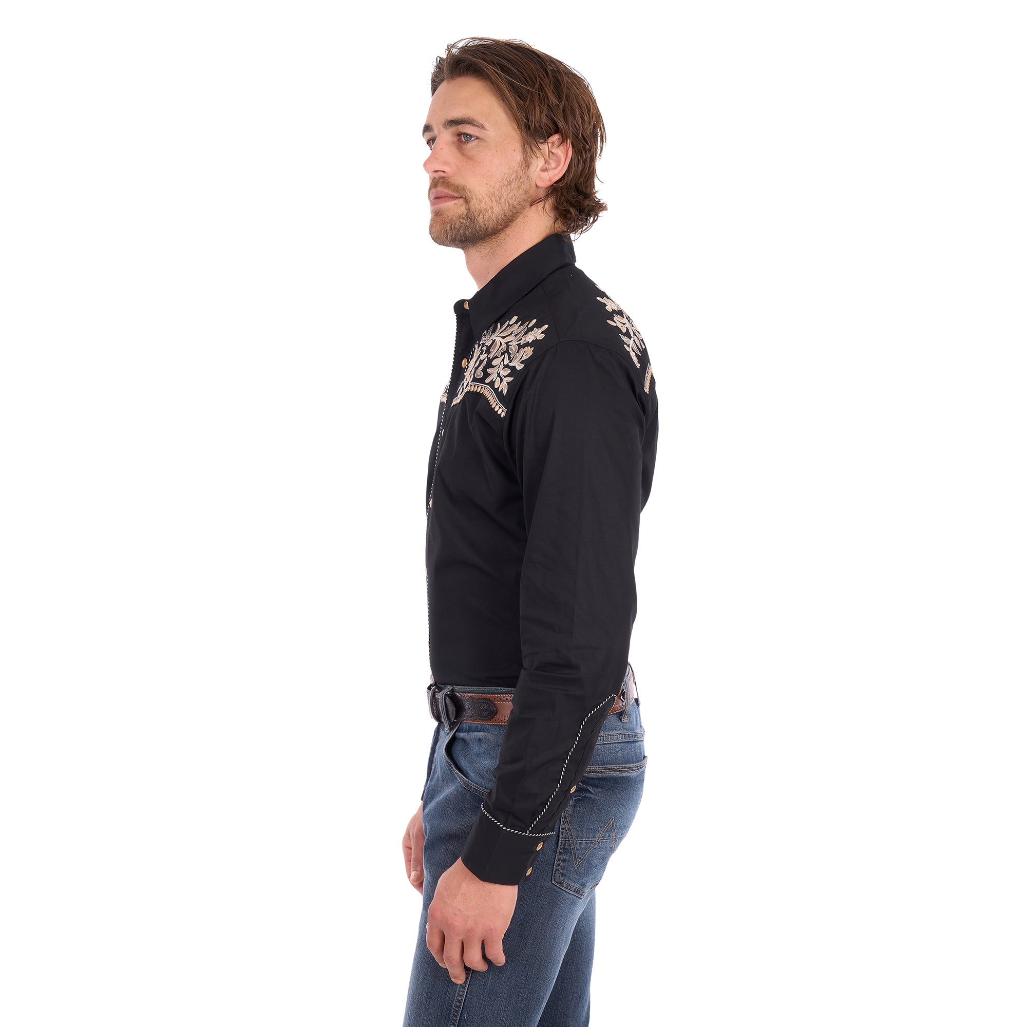 A man wearing a black Wrangler Men's Anthony Embroidered L/S Shirt (X6S1113674) Black with intricate white floral embroidery across the chest, paired with blue jeans and a brown belt, against a plain white background.