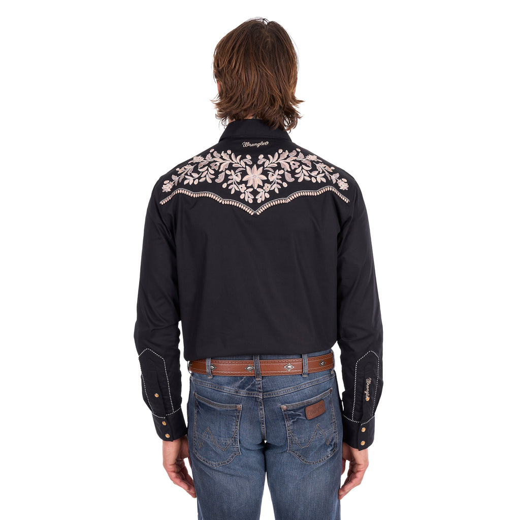 A man wearing a black Wrangler Men's Anthony Embroidered L/S Shirt (X6S1113674) Black with intricate white floral embroidery across the back, paired with blue jeans and a brown belt, against a plain white background.