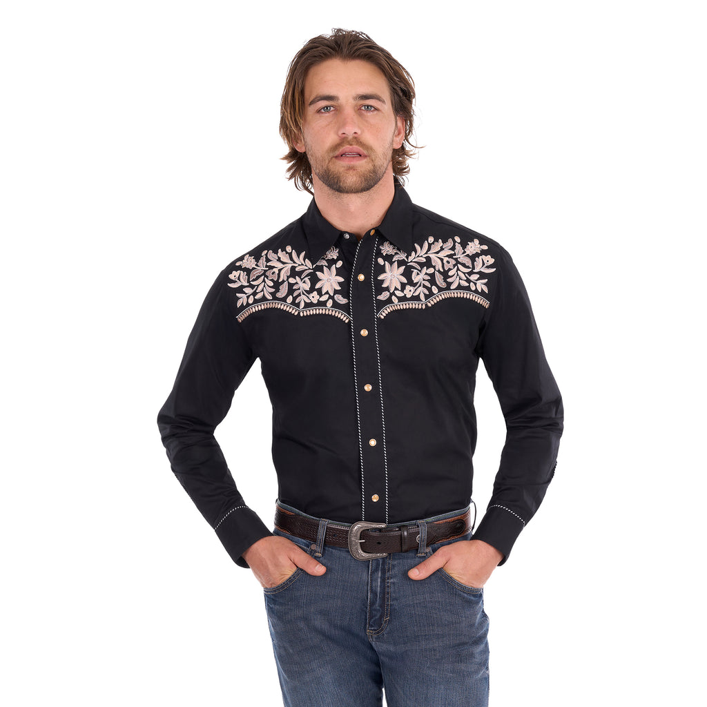 A man wearing a black Wrangler Men's Anthony Embroidered L/S Shirt (X6S1113674) Black with intricate white floral embroidery across the chest, paired with blue jeans and a brown belt, against a plain white background.