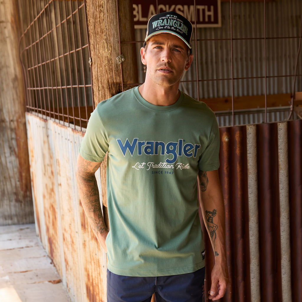 A man wearing a sage green Wrangler Men's Charlie Short Sleeve Tee (X6S1557697) with blue Wrangler logo on the chest, paired with blue jeans, standing in a rustic corrugated metal and wood shed.