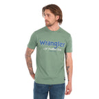 A man wearing a sage green Wrangler Men's Charlie Short Sleeve Tee (X6S1557697) with blue Wrangler logo on the chest, paired with blue jeans, set against a plain white background.