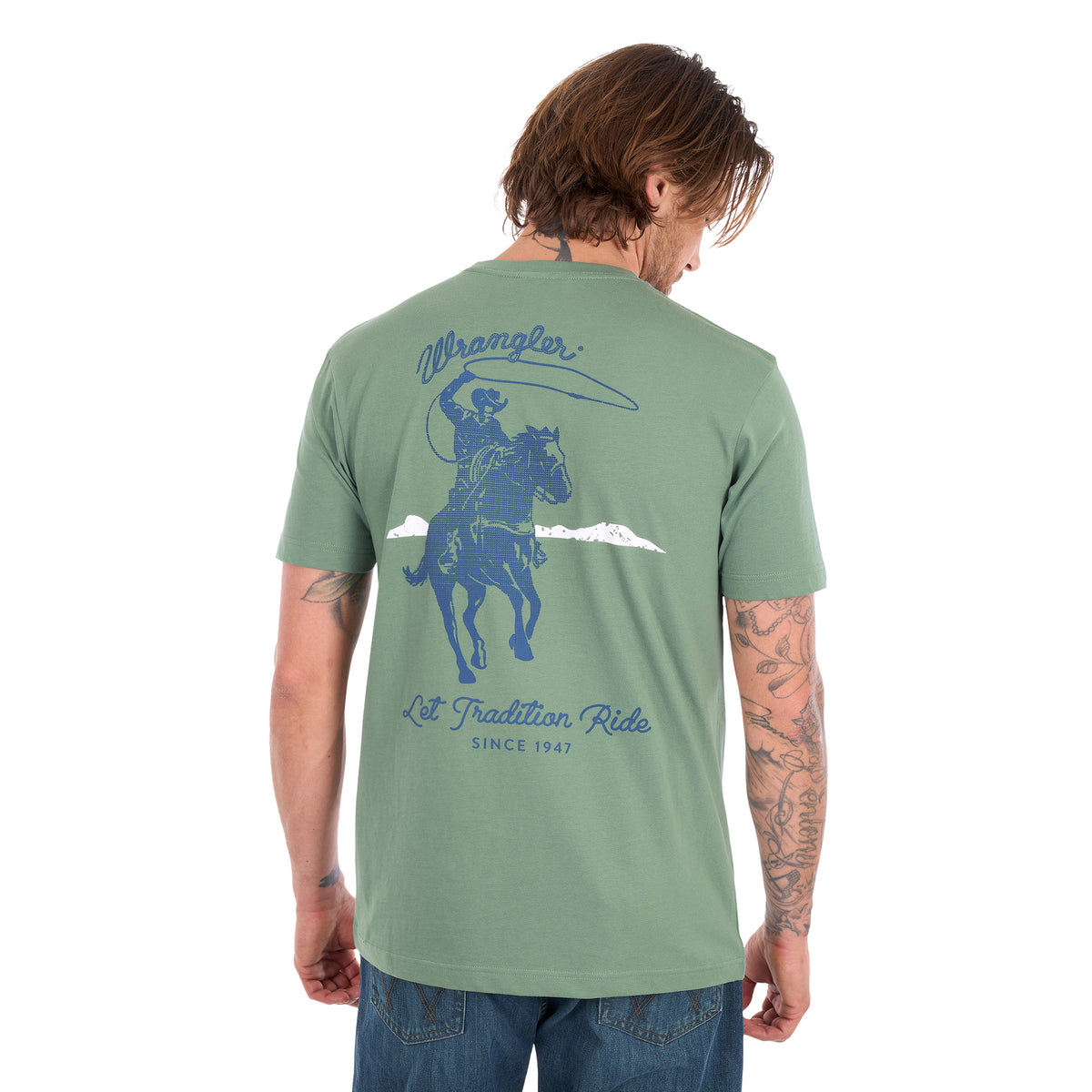 A man wearing a sage green Wrangler Men's Charlie Short Sleeve T-Shirt (X6S1557697), viewed from the back. The shirt features a blue graphic of a cowboy on a horse with a lasso and the text 'Wrangler' and 'Let Tradition Ride' SINCE 1947'.