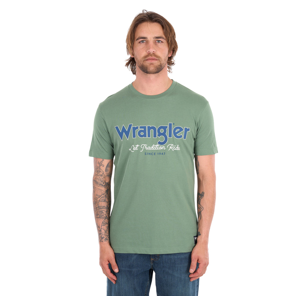 A man wearing a sage green Wrangler Men's Charlie Short Sleeve Tee (X6S1557697) with blue Wrangler logo on the chest, paired with blue jeans, set against a plain white background.