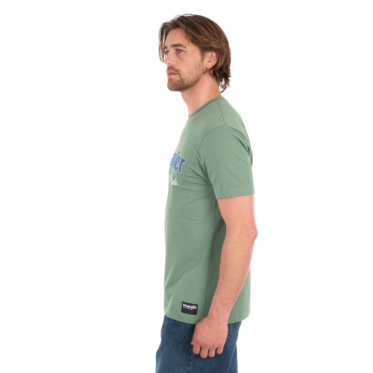 A man in profile wearing a sage green Wrangler Men's Charlie Short Sleeve Tee (X6S1557697) with blue Wrangler logo on the chest, paired with blue jeans, set against a plain white background.