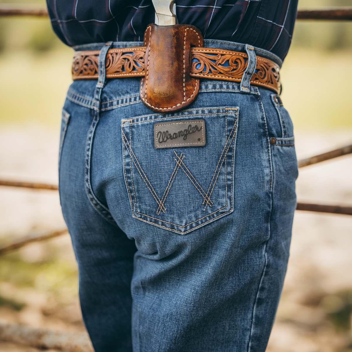 A close up image of a man modelling a pair of Blue Stone Wrangler Men's Cody Johnson 01 Competition Relaxed Fit Jeans (112369005) paired with a western belt that features a knife sheath with pocket knife, and a grey shirt, shown in front of a gate in a rural setting.