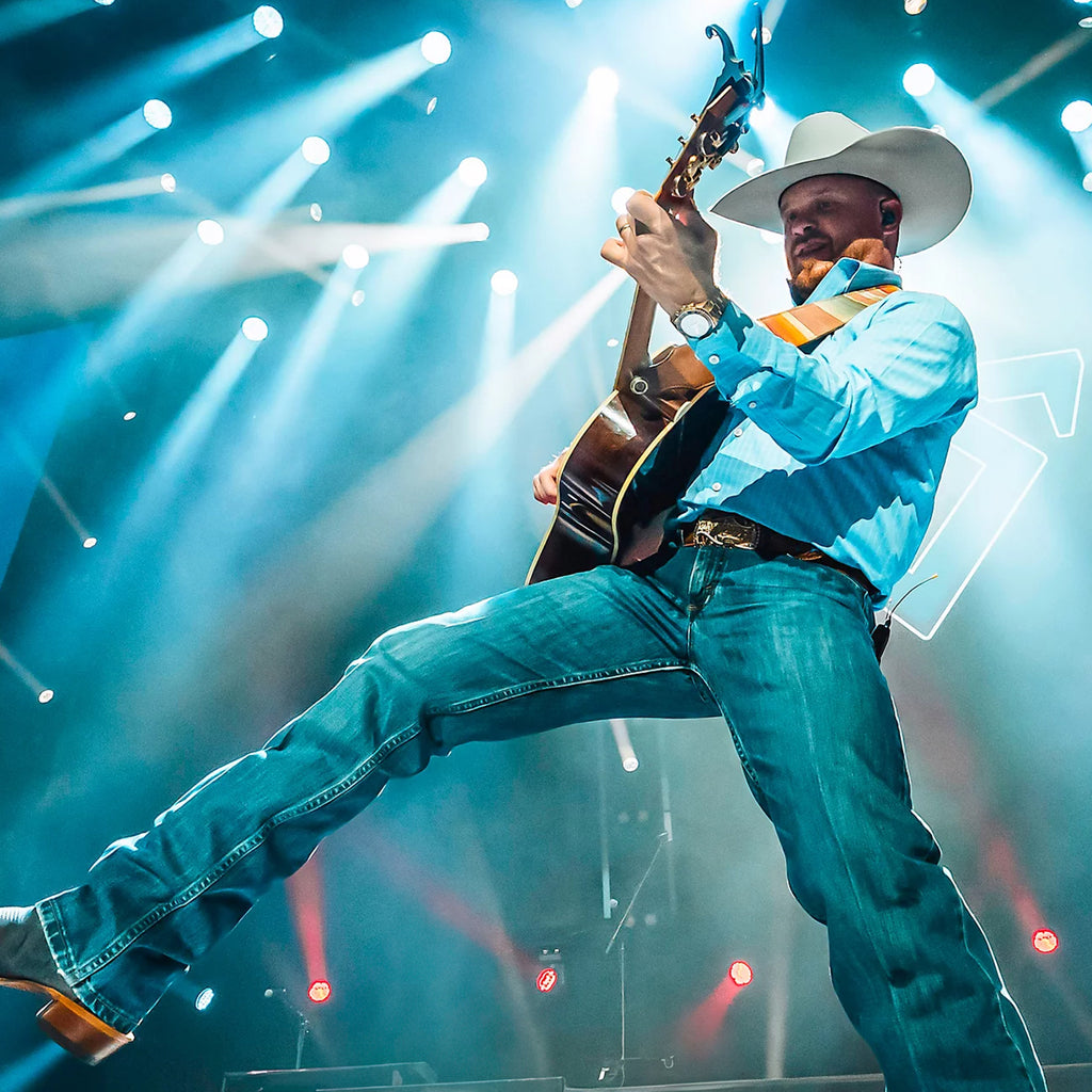 Cody Johnson modelling a pair of Blue Stone Wrangler Men's Cody Johnson 01 Competition Relaxed Fit Jeans (112369005) paired with western boots, a western belt, a blue shirt and a western hat. Shown on stage playing a guitar under bright lights.