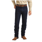 A person stands against a white background, showing only their lower half wearing Wrangler Mens Cowboy Cut Active Flex Jeans (13MAFPW36) in Prewashed Indigo. They're also dressed in a beige shirt, brown belt, and brown cowboy boots.