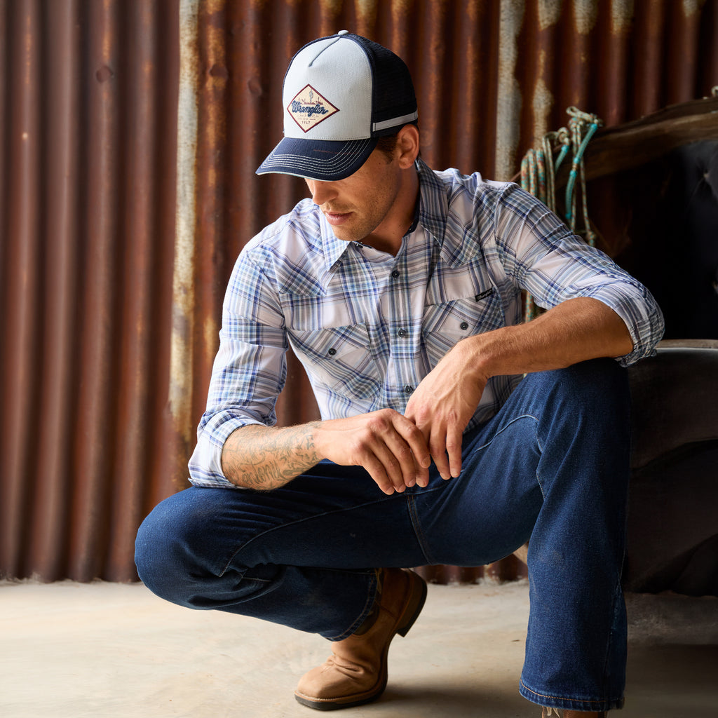 A man crouching in a rustic setting wearing a light blue, royal blue and white Wrangler Men's Eric long-sleeve western-style plaid button-down Shirt (X6S1111667) paired with dark blue jeans and a matching Wrangler Hank High Profile Trucker Cap (X6S1943CAP), set against a plain white background.