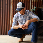 A man crouching in a rustic setting wearing a light blue, royal blue and white Wrangler Men's Eric long-sleeve western-style plaid button-down Shirt (X6S1111667) paired with dark blue jeans and a matching Wrangler Hank High Profile Trucker Cap (X6S1943CAP), set against a plain white background.