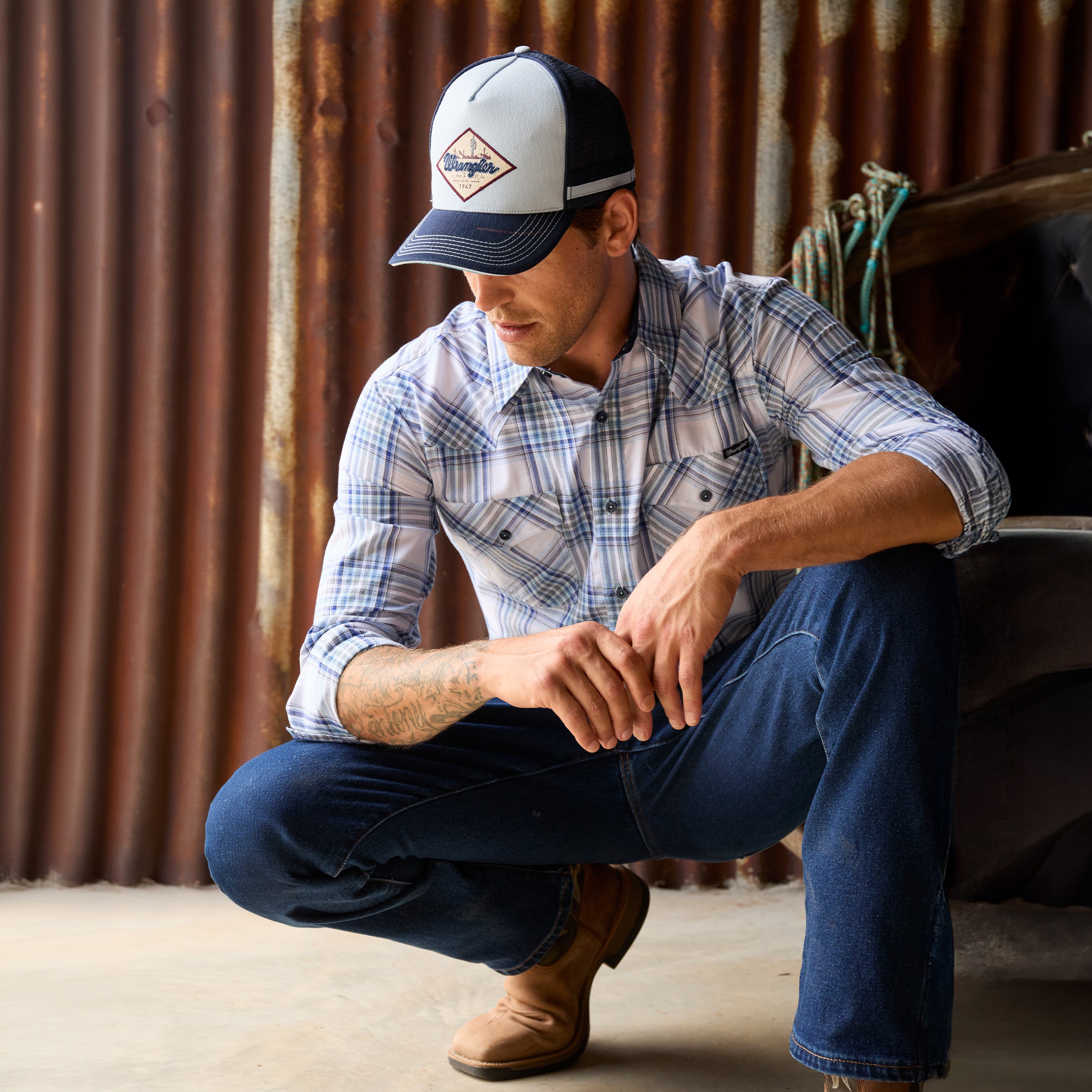 A man crouching in a rustic setting wearing a light blue, royal blue and white Wrangler Men's Eric long-sleeve western-style plaid button-down Shirt (X6S1111667) paired with dark blue jeans and a matching Wrangler Hank High Profile Trucker Cap (X6S1943CAP), set against a plain white background.