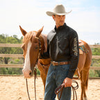 A man wearing a black Wrangler Men's Flex Logo Long Sleeve Shirt (XCP1155359) with a light tan Wrangler Benson Belt (XCP1907BLT) and blue jeans, stands beside a brown horse in an outdoor ranch setting, holding the horses reins.