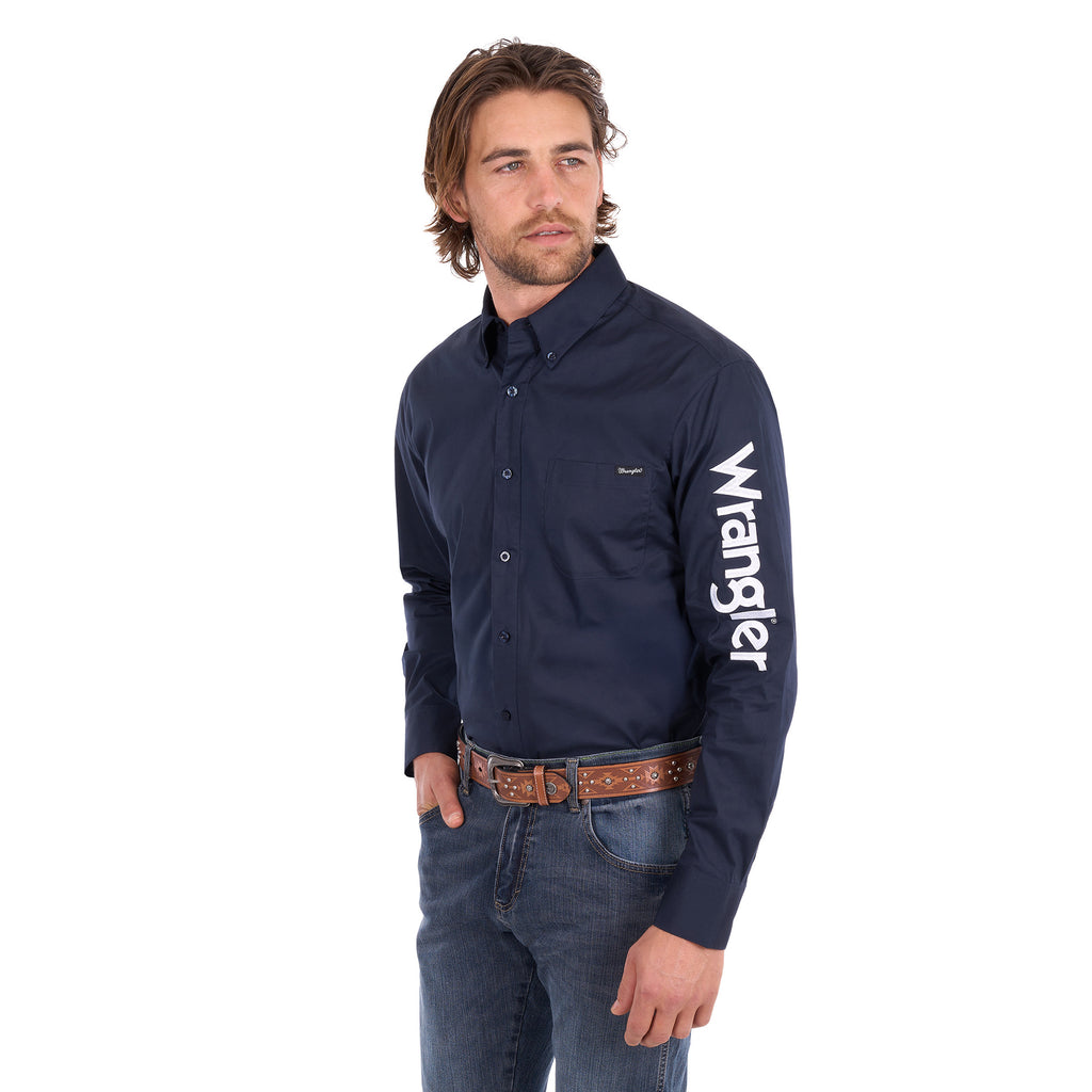 A man wearing a navy blue Wrangler Men's Flex Logo Long Sleeve Shirt (XCP1155359) with a white logo printed vertically on the left sleeve, paired with blue jeans and a Wrangler Onyx Belt (X6S1969BLT), against a plain white background.