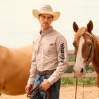 A man wearing a sand Wrangler Men's Flex Logo Long Sleeve Shirt (XCP1155359) with a black logo printed vertically on the left sleeve, paired with blue jeans, a Wrangler Leonard Belt (X6S1909BLT), and a cowboy hat stands in a sandy outdoor arena next to a brown horse.