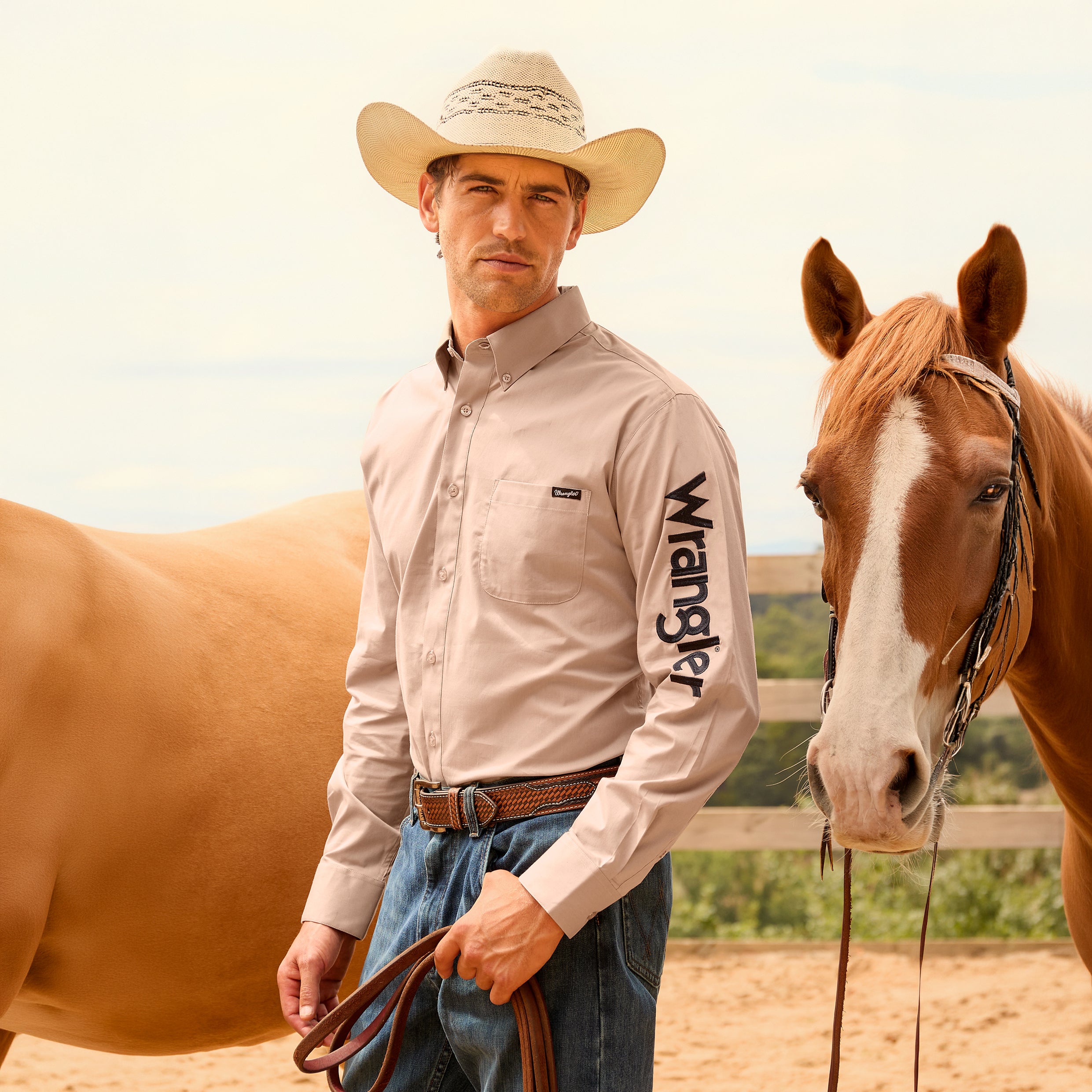A man wearing a sand Wrangler Men's Flex Logo Long Sleeve Shirt (XCP1155359) with a black logo printed vertically on the left sleeve, paired with blue jeans, a Wrangler Leonard Belt (X6S1909BLT), and a cowboy hat stands in a sandy outdoor arena next to a brown horse.