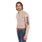 A man wearing a sand Wrangler Men's Flex Logo Long Sleeve Shirt (XCP1155359) with a black logo printed vertically on the left sleeve, paired with blue jeans and a Wrangler Onyx Belt (X6S1969BLT), against a solid white background.
