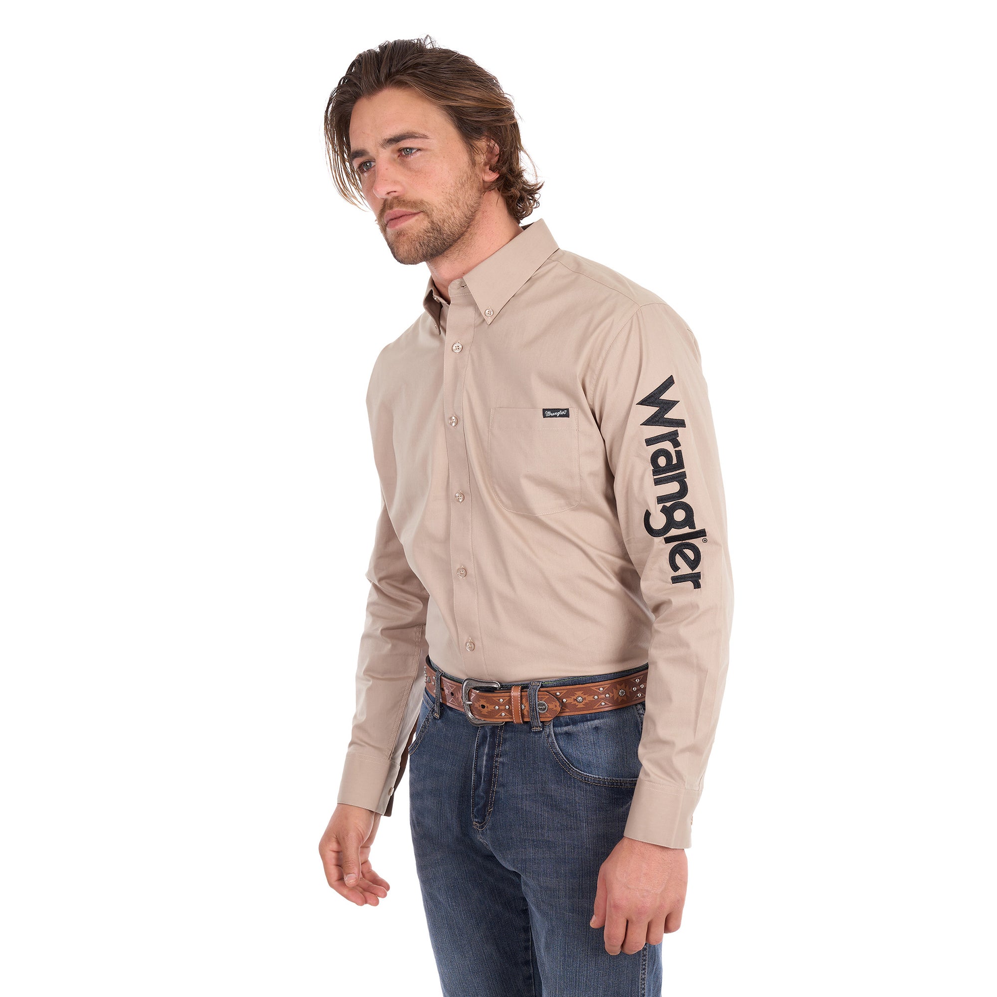 A man wearing a sand Wrangler Men's Flex Logo Long Sleeve Shirt (XCP1155359) with a black logo printed vertically on the left sleeve, paired with blue jeans and a Wrangler Onyx Belt (X6S1969BLT), against a solid white background.