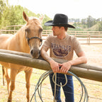 A man wearing a black Wrangler Landon Hat (XCP1955HAT) and a tan Wrangler Men's James Short Sleeve T-Shirt (XCP1557686) with a vintage-style graphic on the chest, paired with blue jeans, leans against a wooden fence next to a light tan horse in an outdoor ranch setting.