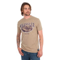 A man wearing a tan Wrangler Men's James Short Sleeve T-Shirt (XCP1557686) with a vintage-style graphic on the chest, paired with blue jeans, photographed against a solid white background.