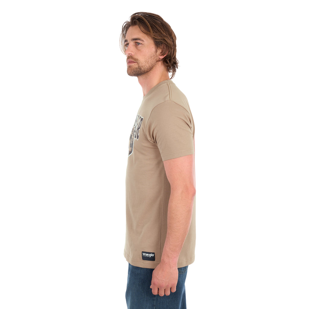 A man in profile wearing a tan Wrangler Men's James Short Sleeve T-Shirt (XCP1557686) with a vintage-style graphic on the chest, paired with blue jeans, photographed against a solid white background.