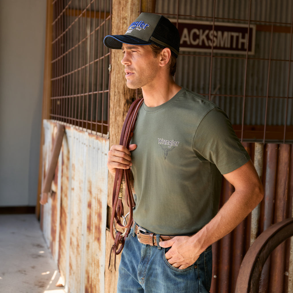 A man wearing a cypress green Wrangler Men's Jay Short Sleeve T-shirt (XCP1557683) with a small Wrangler logo on the left chest, paired with a Wrangler Dave High Profile Trucker Cap (X6S1942CAP) and blue jeans, standing in a rustic barn holding a brown leather bridle.