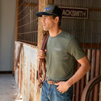 A man wearing a cypress green Wrangler Men's Jay Short Sleeve T-shirt (XCP1557683) with a small Wrangler logo on the left chest, paired with a Wrangler Dave High Profile Trucker Cap (X6S1942CAP) and blue jeans, standing in a rustic barn holding a brown leather bridle.