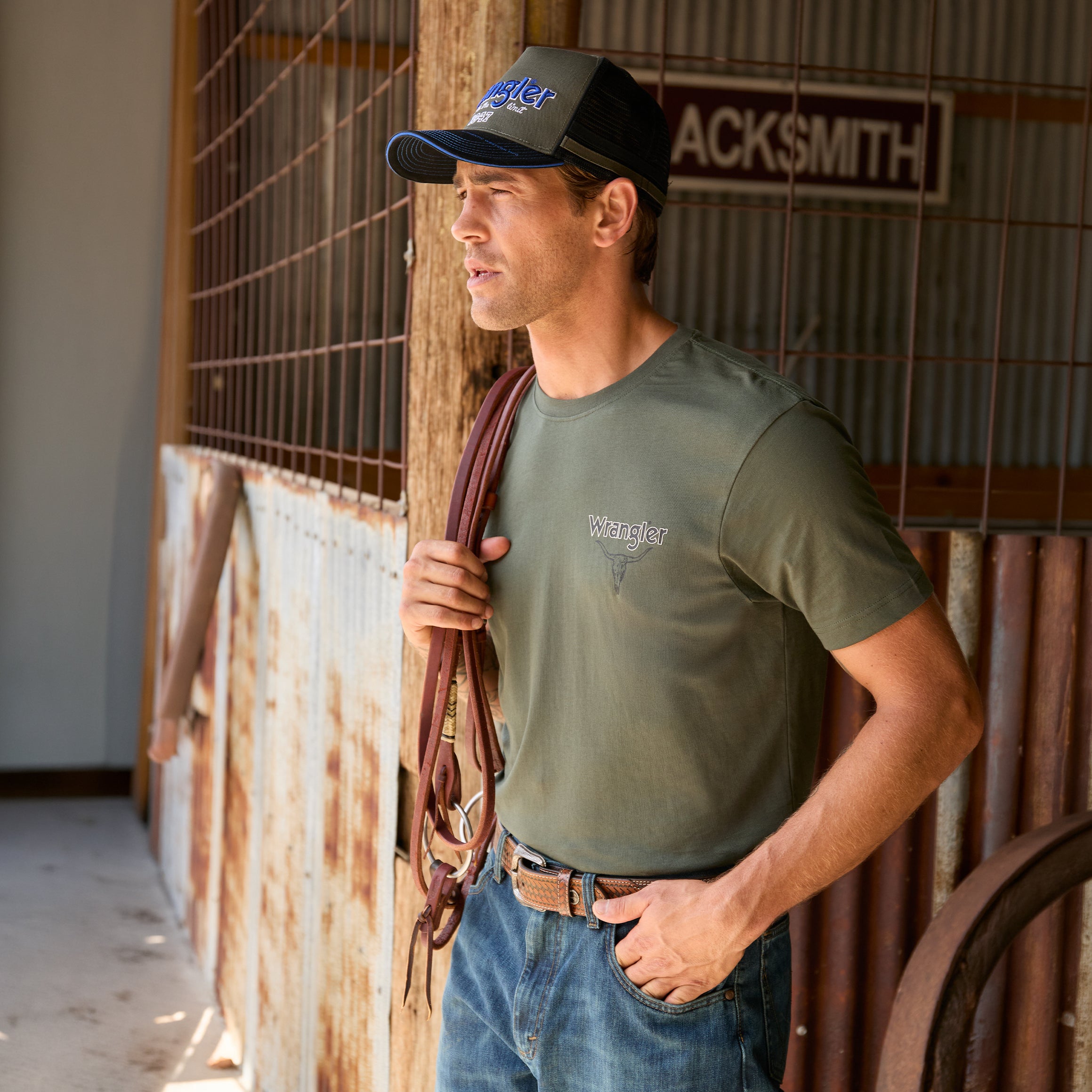 A man wearing a cypress green Wrangler Men's Jay Short Sleeve T-shirt (XCP1557683) with a small Wrangler logo on the left chest, paired with a Wrangler Dave High Profile Trucker Cap (X6S1942CAP) and blue jeans, standing in a rustic barn holding a brown leather bridle.