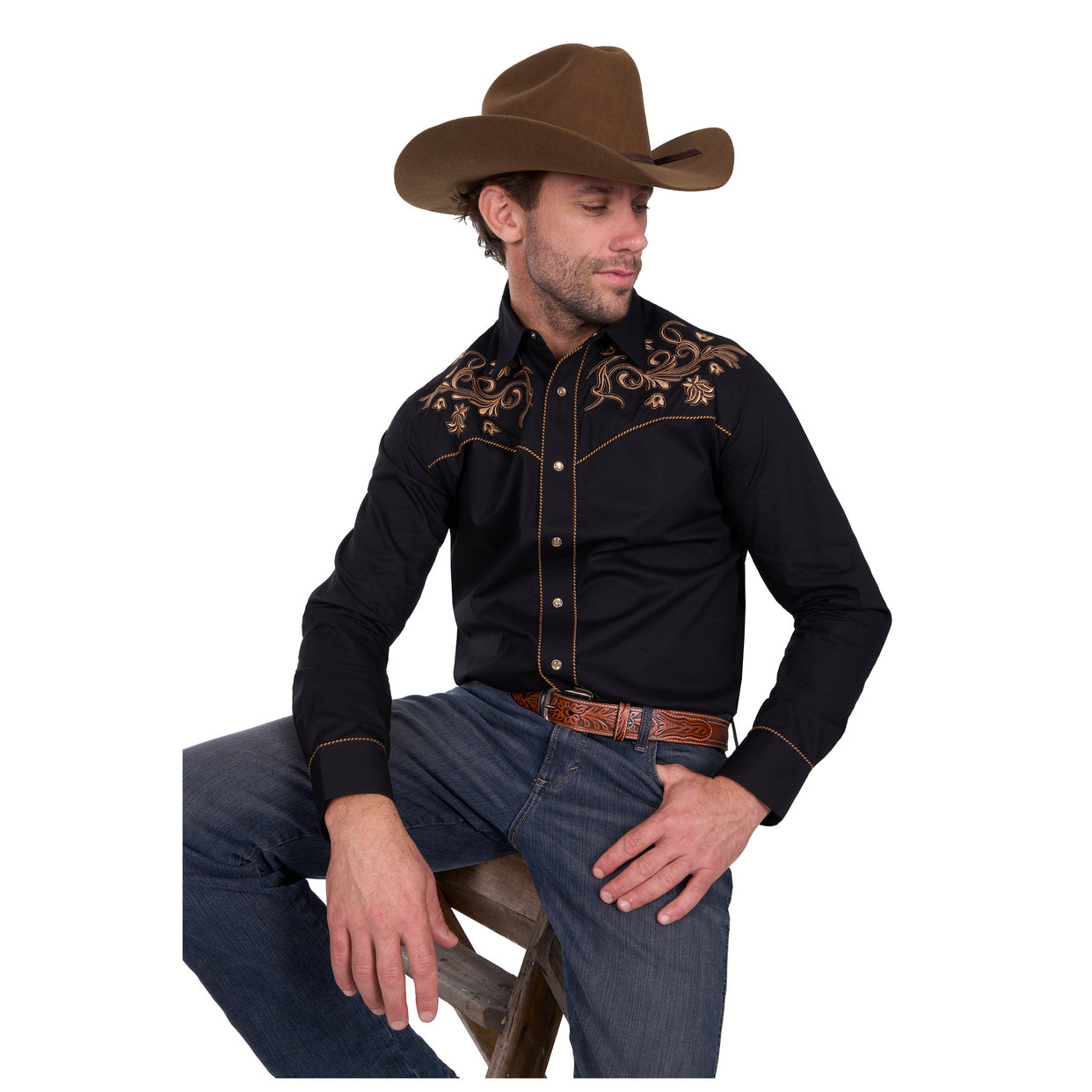 A man wearing a brown cowboy hat, Wrangler Mens Luke Embroided L/S Shirt (X6W1111494) Black, blue jeans, and a patterned belt sits on a wooden stool, looking down and to the side.