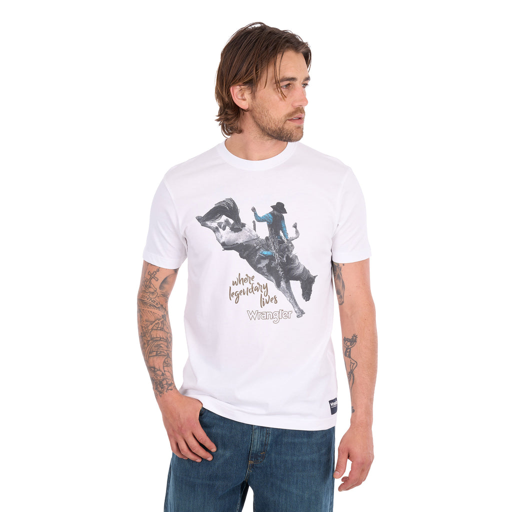 A man wearing a white Wrangler Men's Michael Short Sleeve Tee (X6S1557684) with a graphic of a cowboy riding a bucking bronco and the text 'where legendary lives' and a Wrangler logo, paired with blue jeans, against a plain white background.