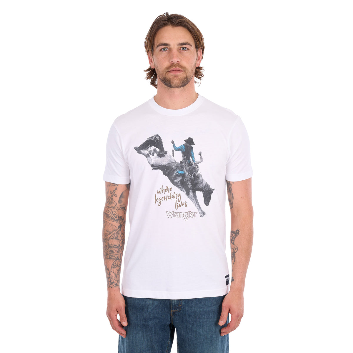 A man wearing a white Wrangler Men's Michael Short Sleeve Tee (X6S1557684) with a graphic of a cowboy riding a bucking bronco and the text 'where legendary lives' and a Wrangler logo, paired with blue jeans, against a plain white background.